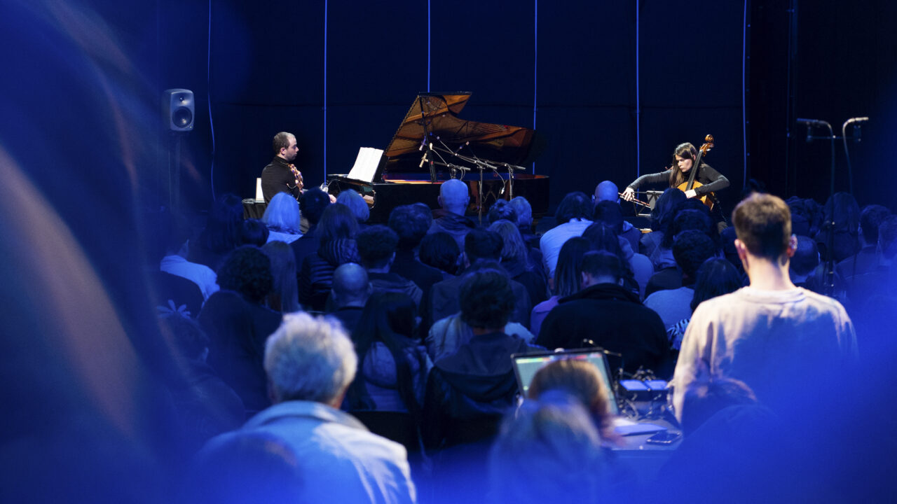 A concert: a pianist and a cellist perform before a focused audience under blue light.