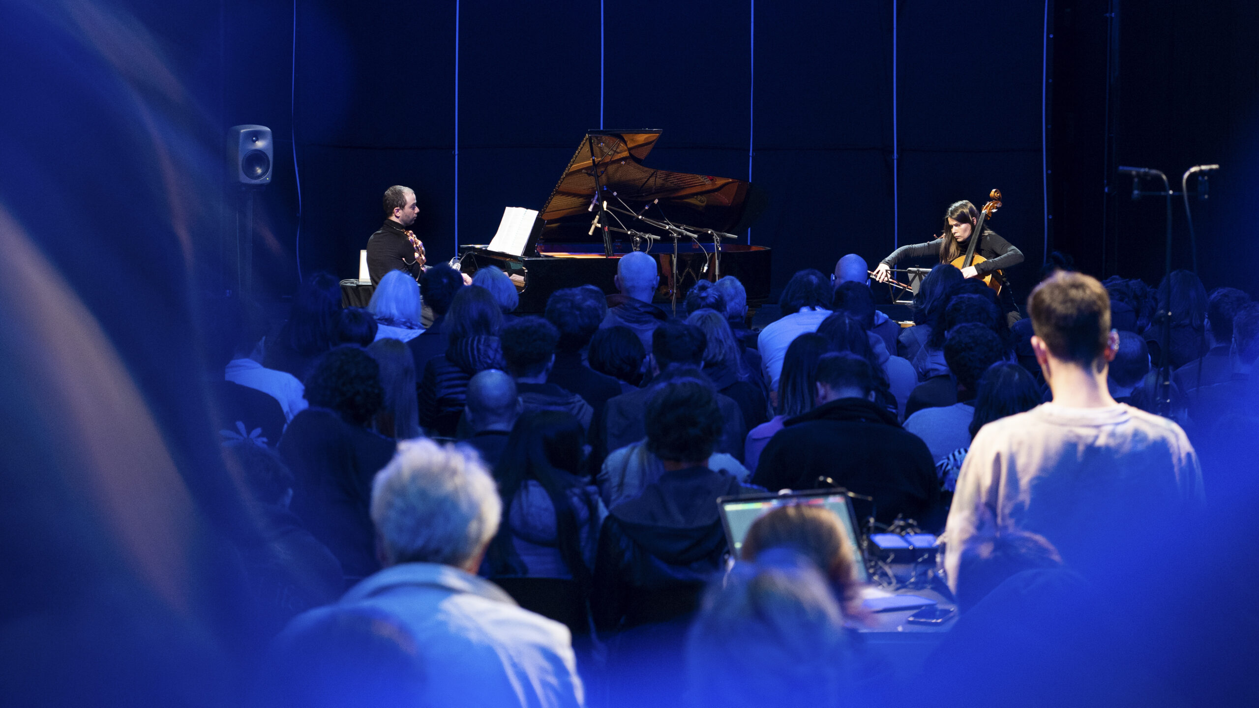 Ein Konzert: Ein Pianist und eine Cellistin spielen vor einem konzentrierten Publikum in blauem Licht.