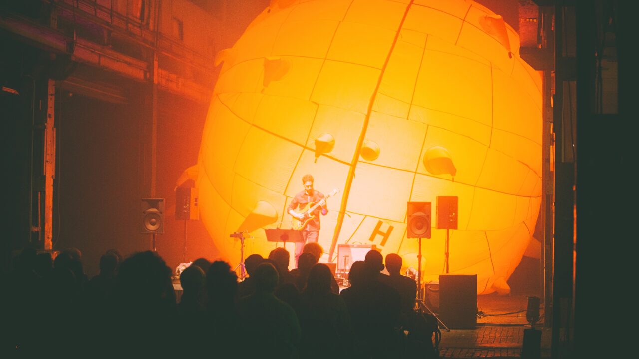 A musician with an electric guitar performs in front of a huge glowing orange balloon-like object, while the audience watches.