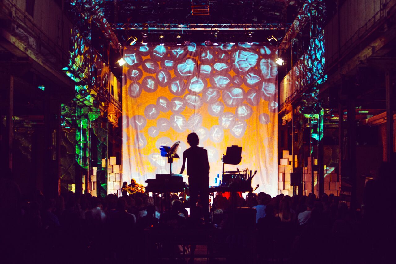 A large hall with colorful light projection on a curtain; in the foreground, the silhouette of a person at a projection table, in the background, musicians on stage.