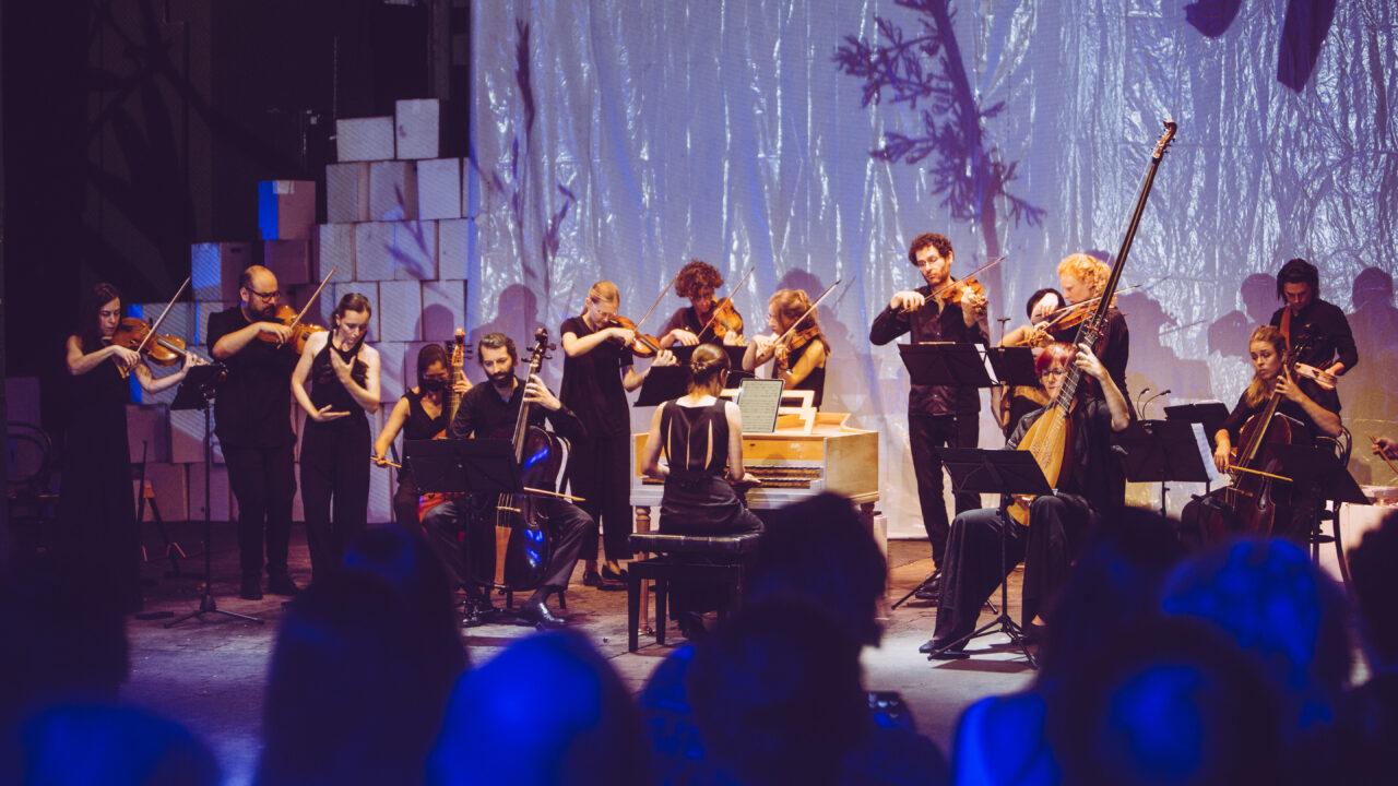 An ensemble of musicians with string instruments and harpsichord performs together on stage before an audience.