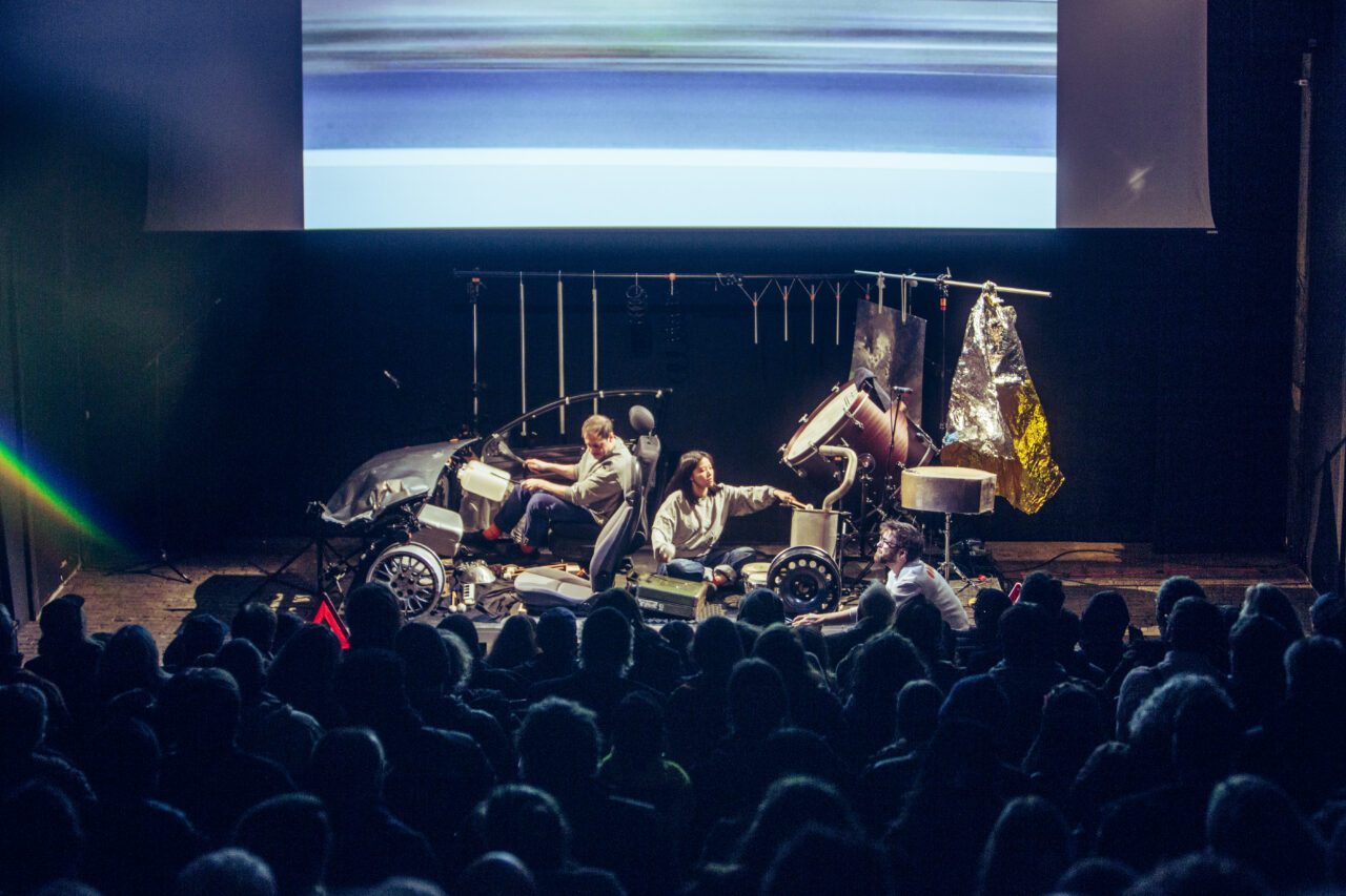 On a stage, an ensemble sits among car parts and metallic objects, while a video is projected in the background. In front of the stage, the audience sits in semi-darkness.