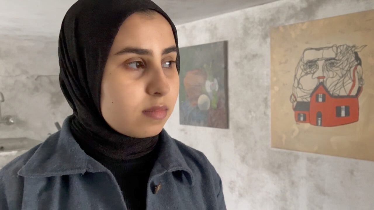 Eine junge Frau mit schwarzem Kopftuch und dunkler Jacke ist im Vordergrund zu sehen. Sie blickt zur Seite. Im Hintergrund befindet sich ein heller, schlicht wirkender Raum mit zwei Kunstwerken: Das rechte zeigt ein rotes Haus vor einer skizzenhaften menschlichen Figur, das linke ist eine abstrakte Darstellung.
