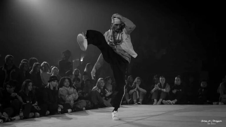 Black-and-white photograph of a dance scene on a stage. In the center, a dancer stands in a dynamic pose: he lifts one leg high forward while the other remains firmly on the ground. One hand rests on his head as part of the choreography. The dancer is wearing light-colored sneakers, dark pants, a light top, and a loosely draped cloth over his head. The background is dark, which strongly highlights the dancer. A spotlight from the upper left illuminates him and creates strong contrasts. Around the stage, audience members sit on the floor and watch the performance attentively.