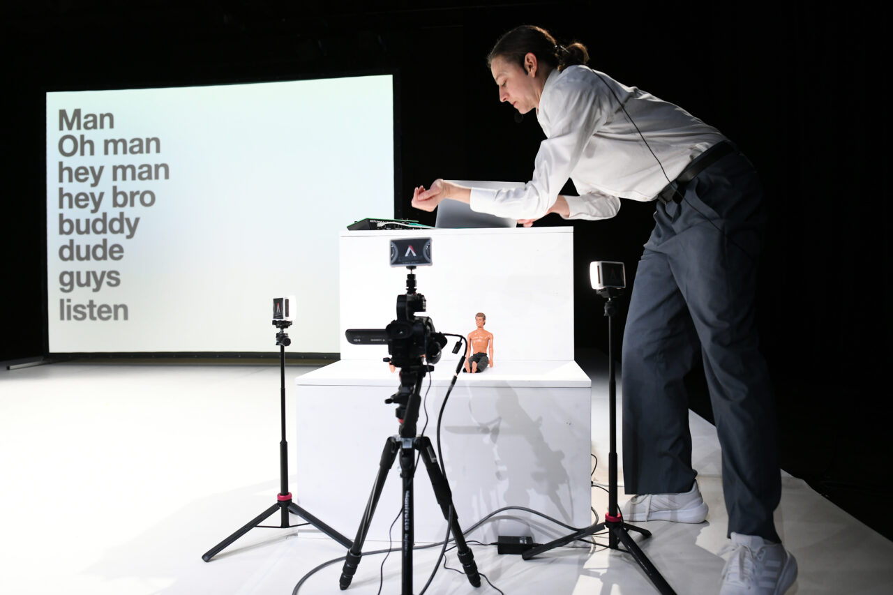 Person in weißem Hemd und grauer Hose arbeitet an einem Tisch mit Puppen, während im Hintergrund groß projizierte Wörter wie ‚Man‘, ‚hey bro‘ und ‚dude‘ zu lesen sind.