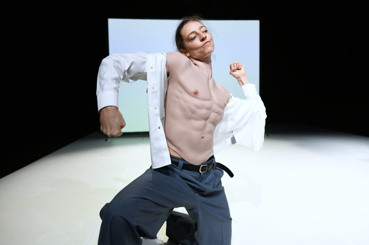 Eine Person im offenen weißen Hemd trägt einen künstlichen Muskelkörper und posiert mit angespannten Armen in einer Macho-Haltung.