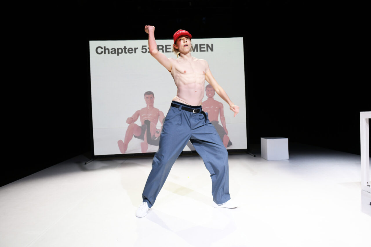 Eine Person mit nacktem Oberkörper trägt einen künstlichen Muskel-Torso, eine rote Baseballkappe und posiert breitbeinig vor einer Projektion mit der Aufschrift ‚Chapter 5: REAL MEN‘