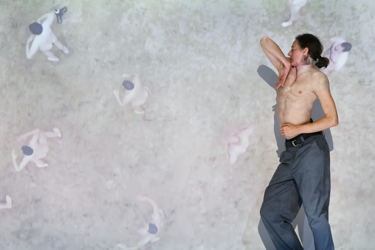 Eine Person mit nacktem Oberkörper und grauer Hose posiert seitlich vor einer Projektion, die nackte Körper von oben zeigt. Der Arm ist elegant angewinkelt, die Pose erinnert an Tanz.