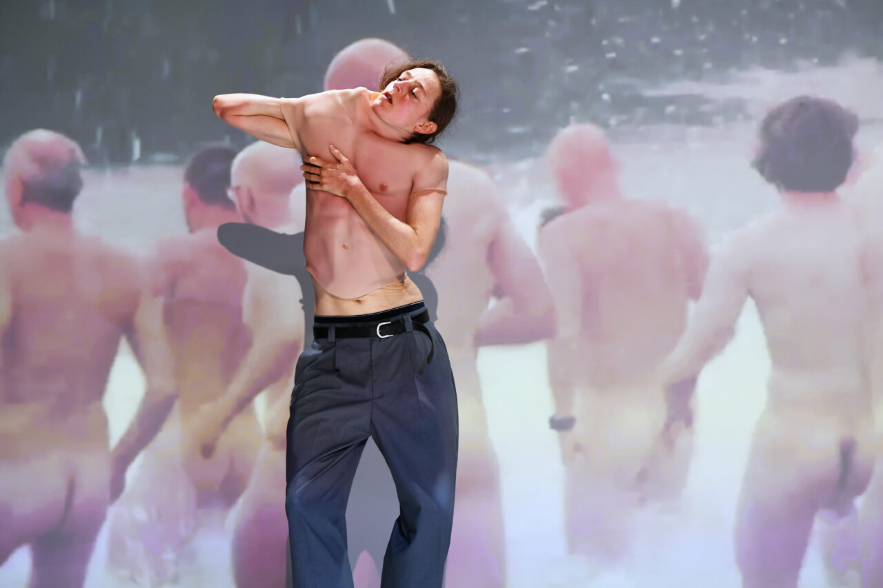 Eine Person im nackten Oberkörper kämpft mit einem hautfarbenen Silikonanzug, während im Hintergrund eine Projektion nackter Männerkörper zu sehen ist.