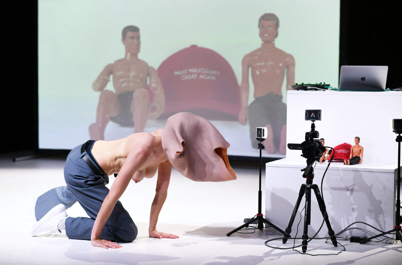 Eine Performer:in kriecht mit nacktem Oberkörper auf allen Vieren, der Kopf steckt in einer umgestülpten Silikonmaske. Hinter ihr sind auf einer Projektion zwei Ken-Puppen und eine rote Kappe mit der Aufschrift ‚Make Masculinity Great Again‘ zu sehen.