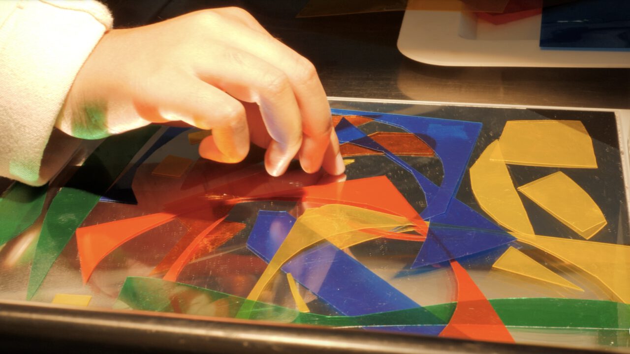 Das Bild zeigt eine Hand, die über eine flache, beleuchtete Oberfläche greift und mehrere transparente, farbige Kunststoffstücke arrangiert. Die Formen sind in kräftigen Farben wie Rot, Blau, Gelb und Grün gehalten und überlappen sich teilweise, wodurch neue Farbmischungen entstehen. Die Szene wirkt kreativ und spielerisch, fast wie ein geometrisches Puzzle oder eine künstlerische Komposition mit Licht und Farbe. Im Hintergrund ist unscharf ein Tisch oder Gerät zu erkennen, während der Fokus klar auf der Hand und den bunten Formen liegt.