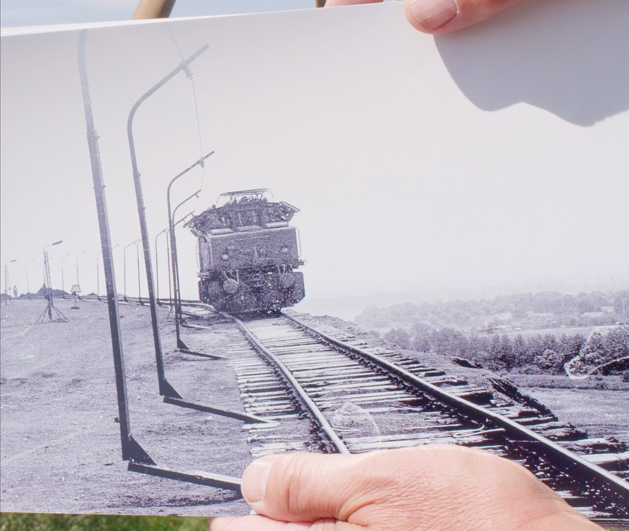Es wird ein altes Schwarz-Weiß-Foto von einer Lokomotive auf Gleisen gezeigt, dass jemanfd in der Hand hält.