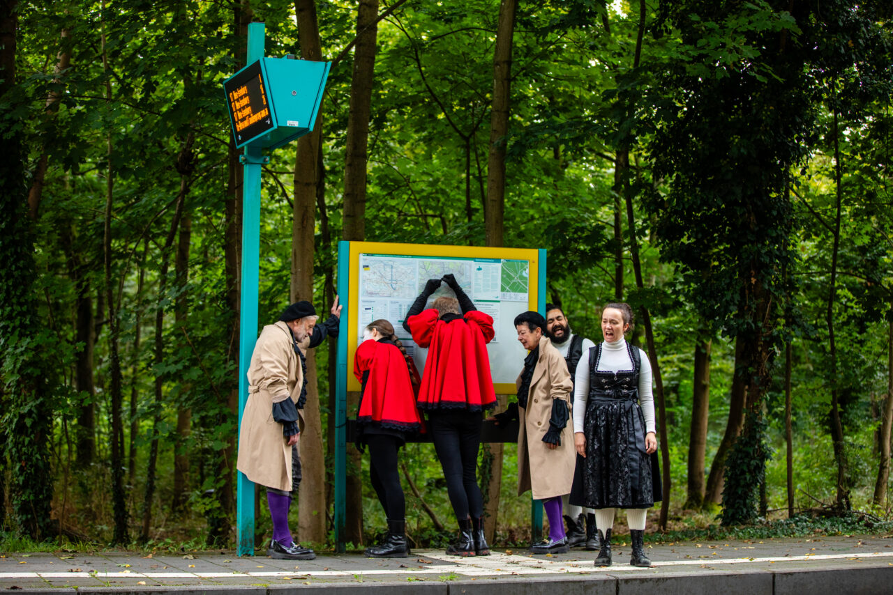 Eine Gruppe von Menschen in auffälligen Kostümen steht an einer Bushaltestelle im Wald vor einem großen Fahrplan.