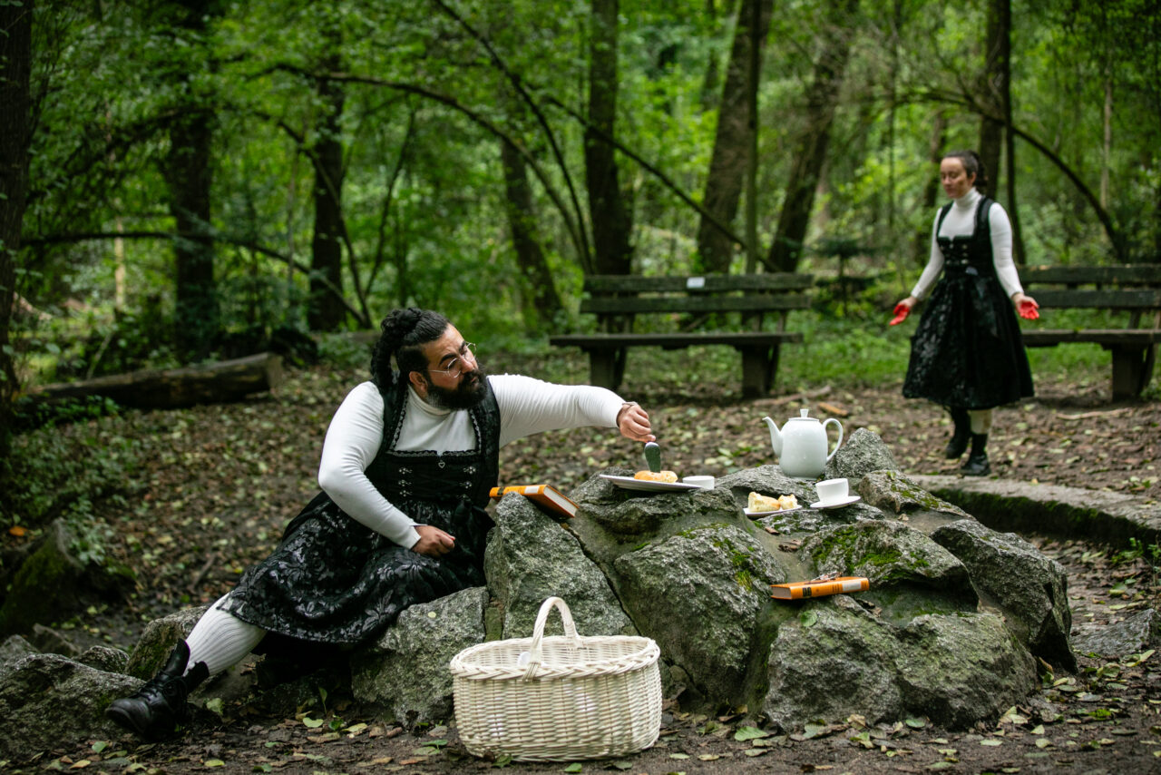 Ein Mann und eine Frau in schwarzen Kleidern mit weißen Blusen inszenieren ein Picknick im Wald mit Teekanne und Kuchen.