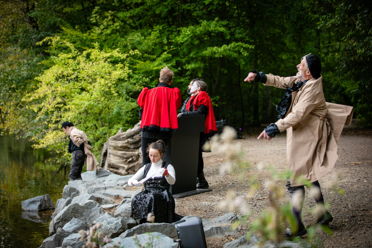 Darsteller:innen in roten Capes und Trenchcoats spielen eine Szene am Ufer eines Waldsees.