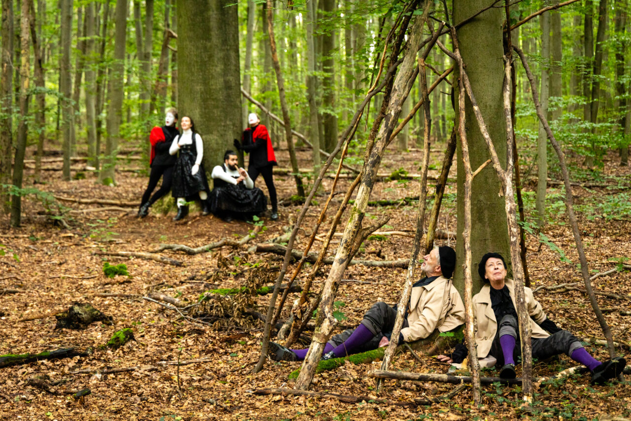 Darsteller:innen im Wald, zwei sitzen unter einer aus Ästen gebauten Struktur, andere stehen und lehnen an Bäumen.“