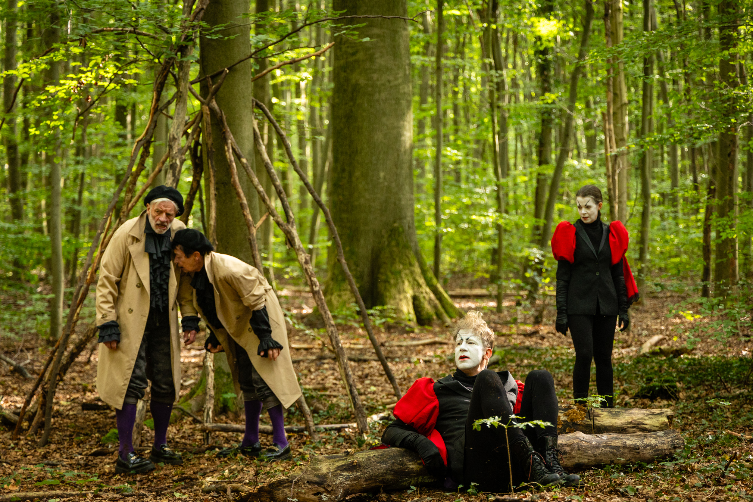 Vier Darsteller:innen im Wald, zwei in Trenchcoats mit lila Strümpfen, zwei mit roten Capes und weiß geschminkten Gesichtern.