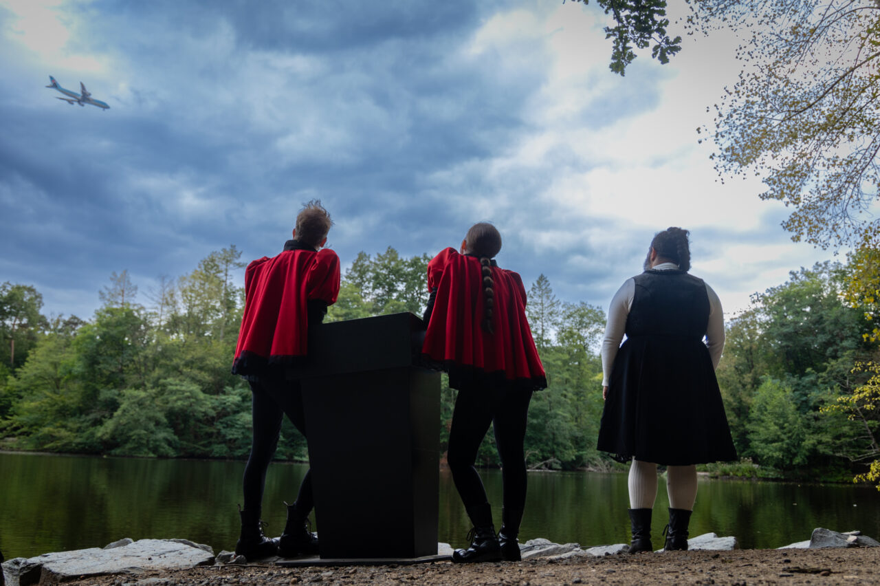 Drei Personen in Kostümen mit roten Umhängen stehen am Ufer eines Sees, über ihnen fliegt ein Flugzeug durch den bewölkten Himmel.