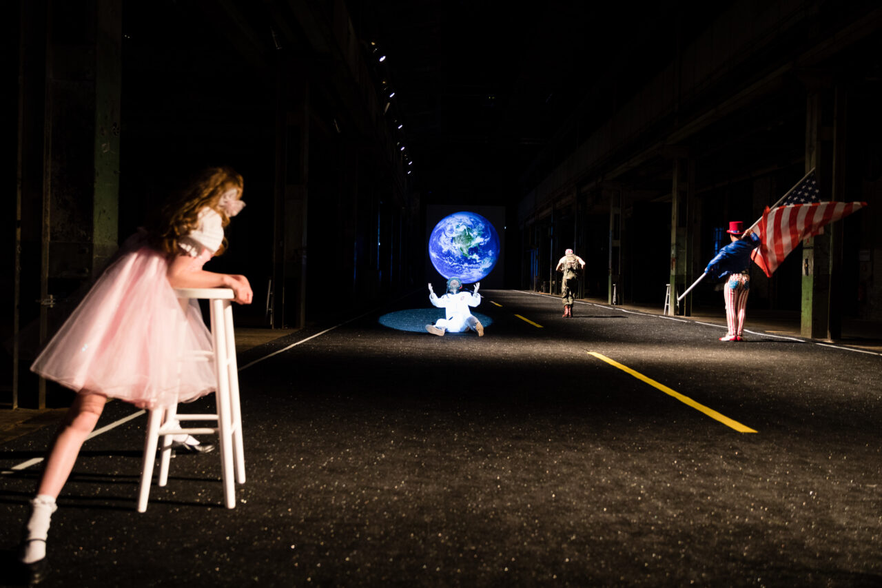 Bühnenbild mit Figuren in Kostümen: eine Frau im rosa Kleid, ein Astronaut, ein Soldat und eine Person mit US-Flagge vor einer Projektion der Erde.