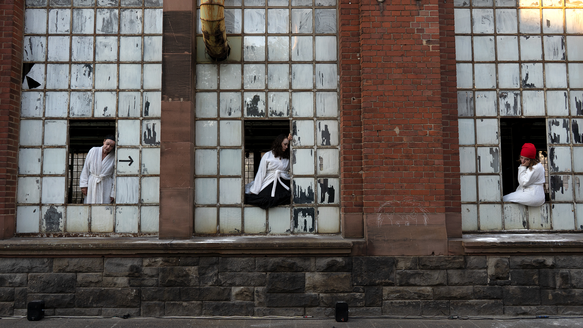 Drei Performer:innen in weißen Bademänteln sitzen oder lehnen in Fensteröffnungen einer alten Backsteinfassade mit Glasbausteinen.