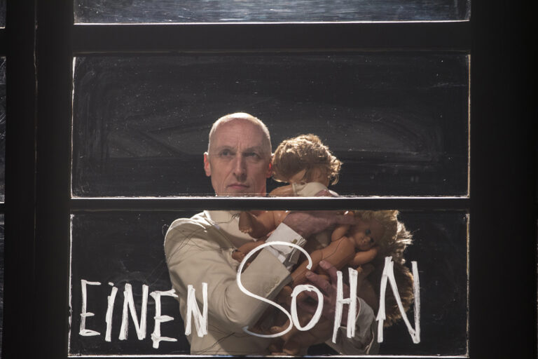 A man in a light suit looks seriously through a window, holding several baby dolls in his arms. On the glass, the words ‘A SON’ are written in white.