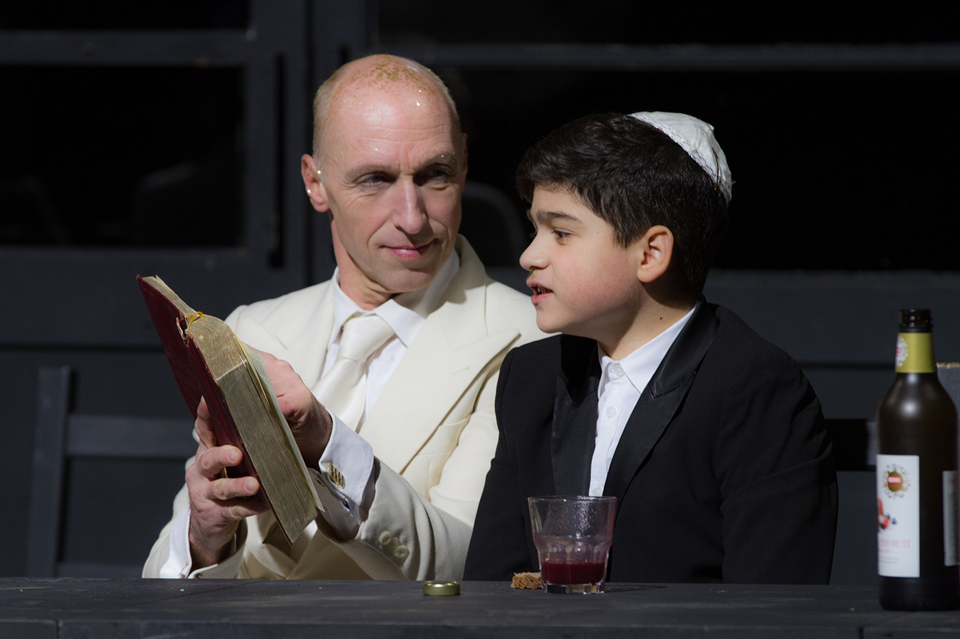 Ein Mann im weißen Anzug sitzt neben einem Jungen mit Kippa. Der Mann hält ein aufgeschlagenes Buch, während der Junge spricht. Auf dem Tisch stehen ein Glas mit roter Flüssigkeit und eine Flasche.
