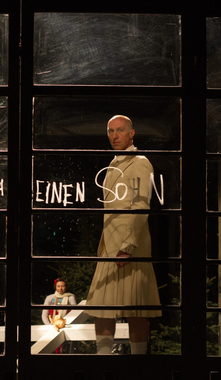 A man in a white suit and pleated skirt stands solemnly in a stage set behind a glass wall with the inscription ‘A SON.’ In the background, a girl with a red ribbon stands beside a white cross.
