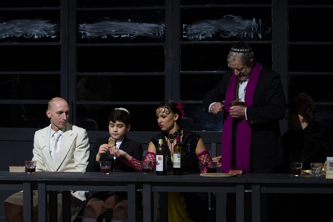 Szenenfoto aus JESUS Weihnachten: Eine festlich gedeckte Tafel mit vier Personen. Links ein Mann im weißen Anzug, daneben ein Junge mit Kippa, eine Frau in glitzerndem Kleid mit Weinflaschen vor sich und rechts ein älterer Mann im schwarzen Anzug mit violettem Schal, der Brot schneidet.