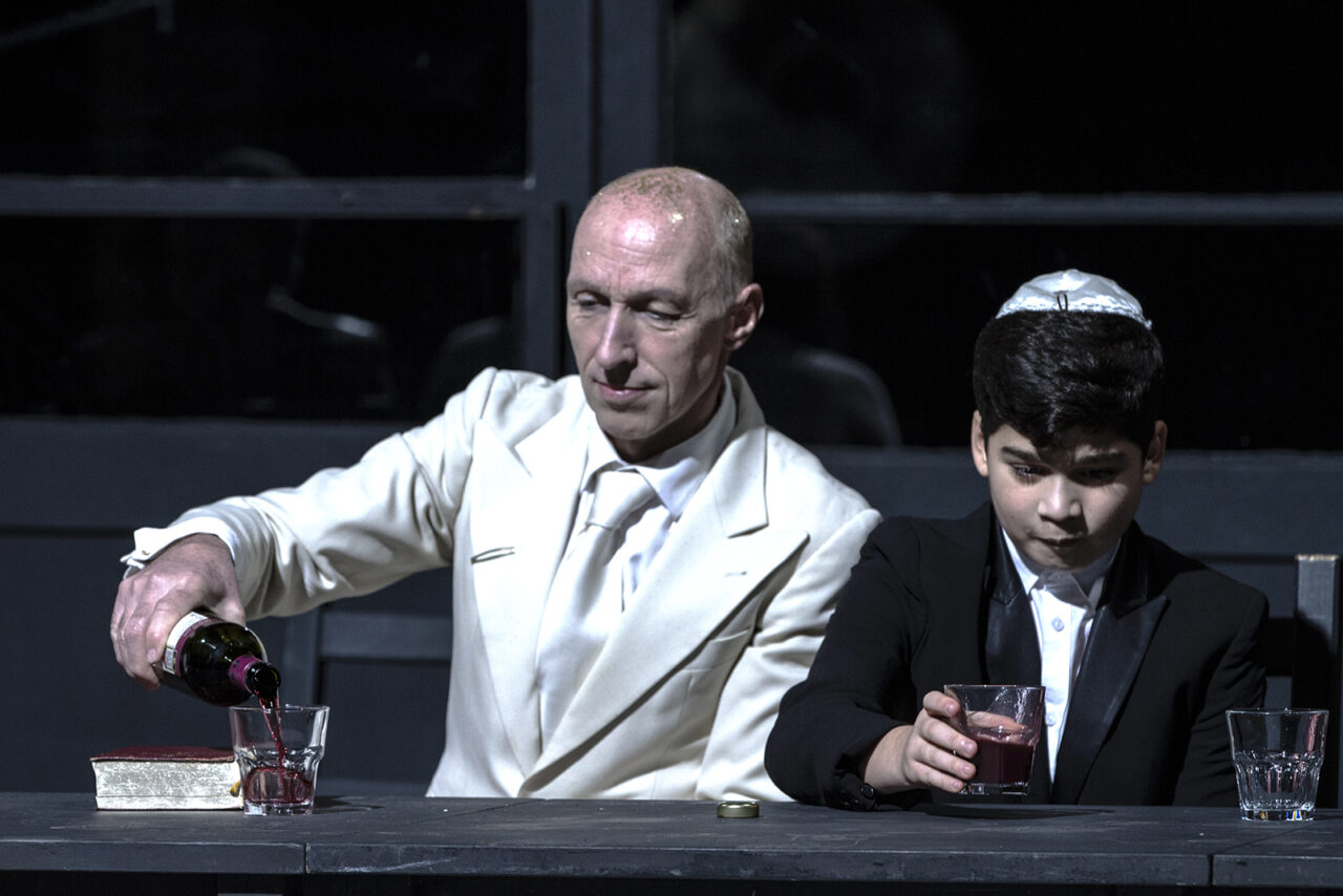Szenenfoto aus JESUS Weihnachten: Ein Mann im weißen Anzug schenkt Rotwein in ein Glas ein, neben ihm sitzt ein Junge im schwarzen Anzug mit Kippa, der konzentriert auf sein eigenes Glas blickt.