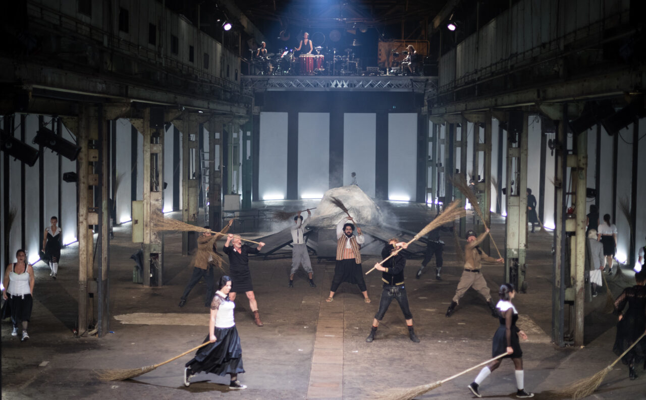 Theaterszene in einer Industriehalle: Viele Performer:innen fegen mit langen Reisigbesen rhythmisch über den Boden, im Hintergrund spielen Musiker:innen auf einer Empore.