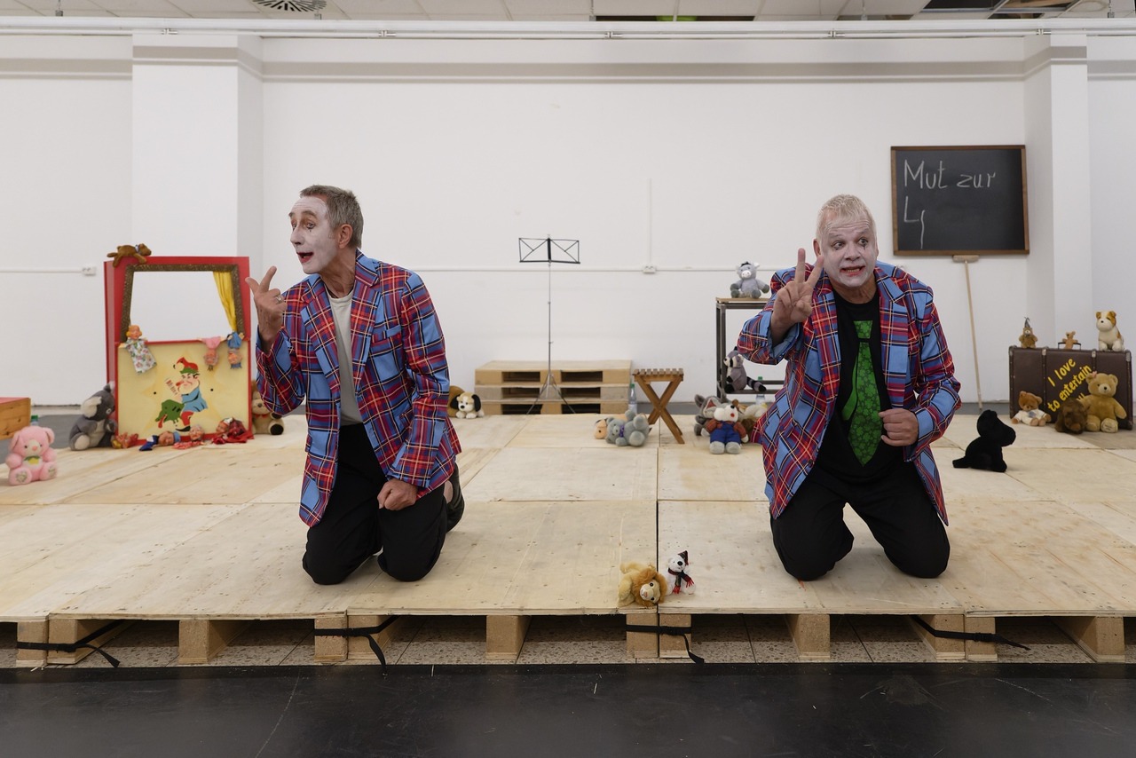 Zwei Performer knien auf einer einfachen Holzpodest-Bühne und tragen auffällige, karierte Sakkos sowie weiß geschminkte Gesichter, die an clowneske Darstellungen erinnern. Mit lebhaften Gesten und ausdrucksstarker Mimik wenden sie sich dem Publikum zu und scheinen in einem dialogischen oder spielerischen Moment zu sein. Die Bühne ist mit Requisiten ausgestattet, darunter Stofftiere, kleine Figuren und eine gerahmte Szenerie, die an ein Puppentheater erinnert. Im Hintergrund steht eine Tafel mit der Aufschrift „Mut zur L…“, was auf ein thematisches Element der Inszenierung hindeutet. Die Szene wirkt bewusst reduziert und zugleich verspielt und erzeugt eine Mischung aus Theater, Performance und kindlich anmutender Fantasiewelt.