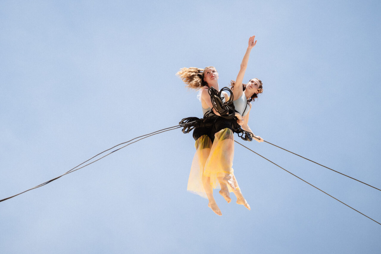Two female acrobats hang secured by ropes high in the air against a blue sky. They wear light tops and yellowish skirts, their bodies intertwined, one stretching an arm upward.