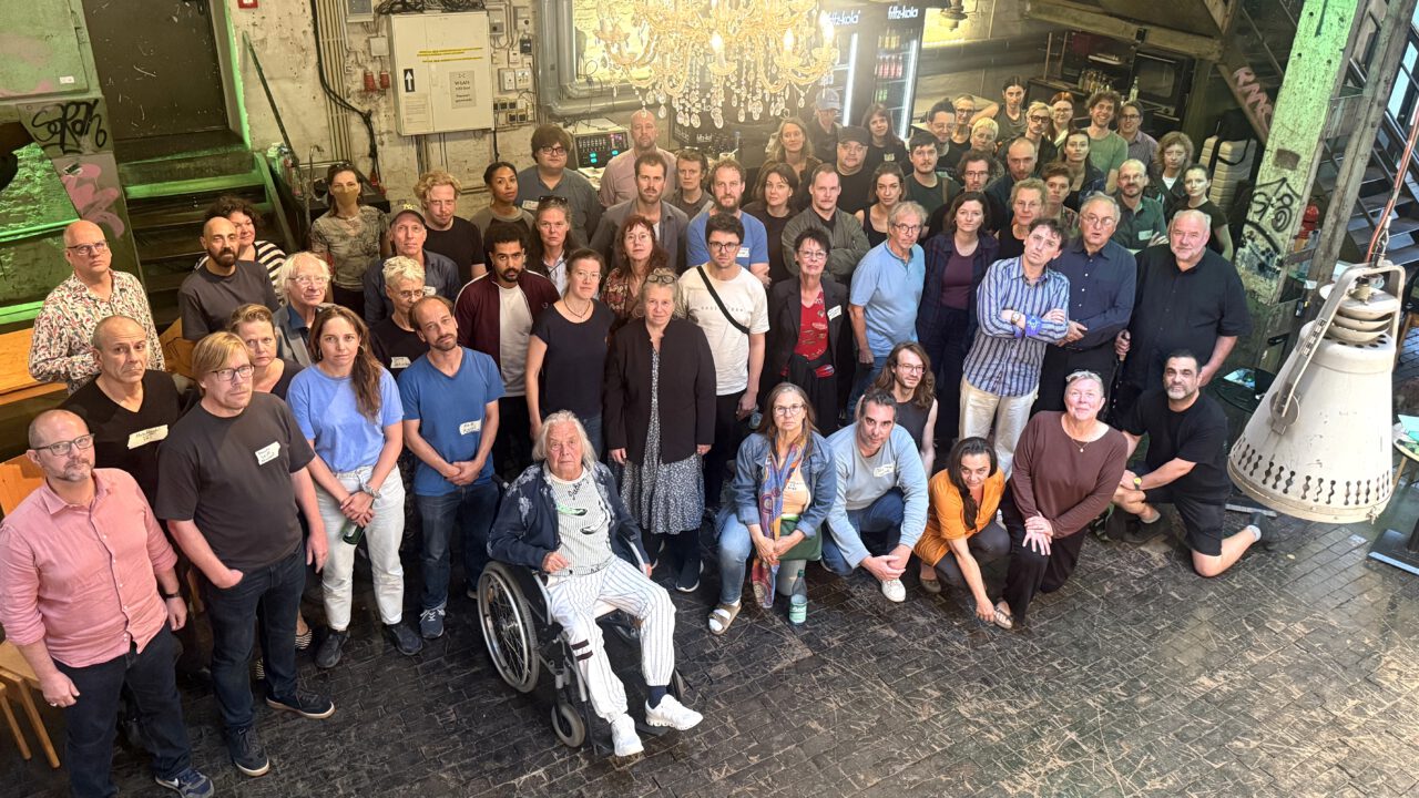 Gruppenfoto der Vollversammlung der Freien Darstellenden Künste Frankfurt im Foyer des Produktionshaus NAXOS. Rund 50 Teilnehmende sind für das gemeinsame Foto versammelt. Im Hintergrund sind das Foyer mit Bar, ein großer Kronleuchter und Backsteinwände zu sehen.