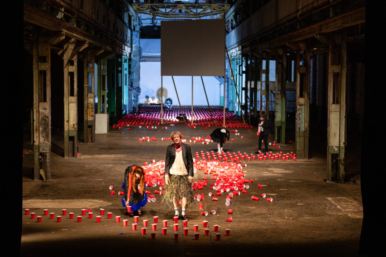 Theaterszene in einer großen Industriehalle mit hunderten roten Plastikbechern am Boden, mehrere Performer:innen in Kostümen bewegen sich zwischen den Bechern.