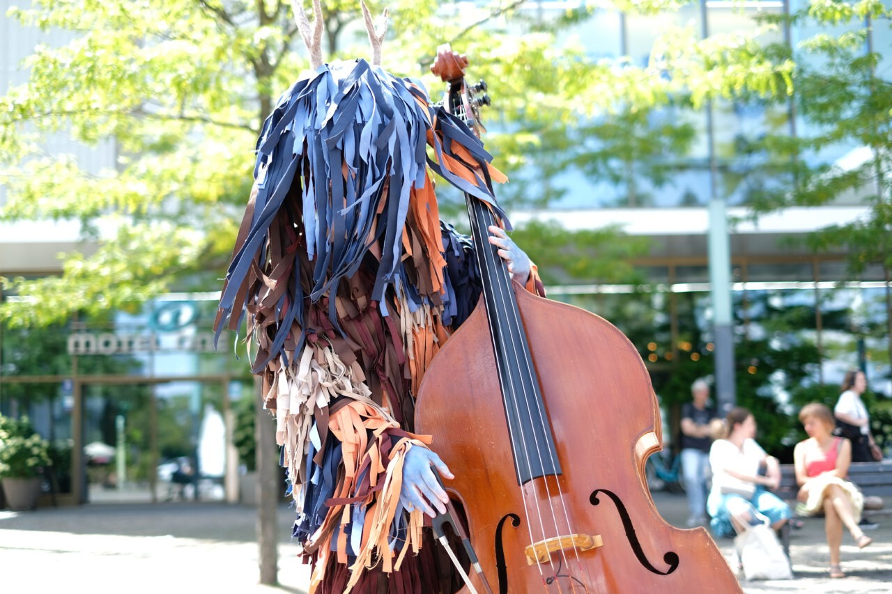 Eine als Wald- oder Fantasiewesen verkleidete Person spielt im Freien auf einem Kontrabass. Das aufwendige Kostüm besteht aus zahlreichen Stoffstreifen in Blau-, Braun- und Beigetönen sowie einem Geweih auf dem Kopf. Die Musikerin oder der Musiker steht auf einem Platz mit Bäumen und moderner Architektur im Hintergrund. Einige Passant*innen sitzen oder bewegen sich im unscharfen Hintergrund.