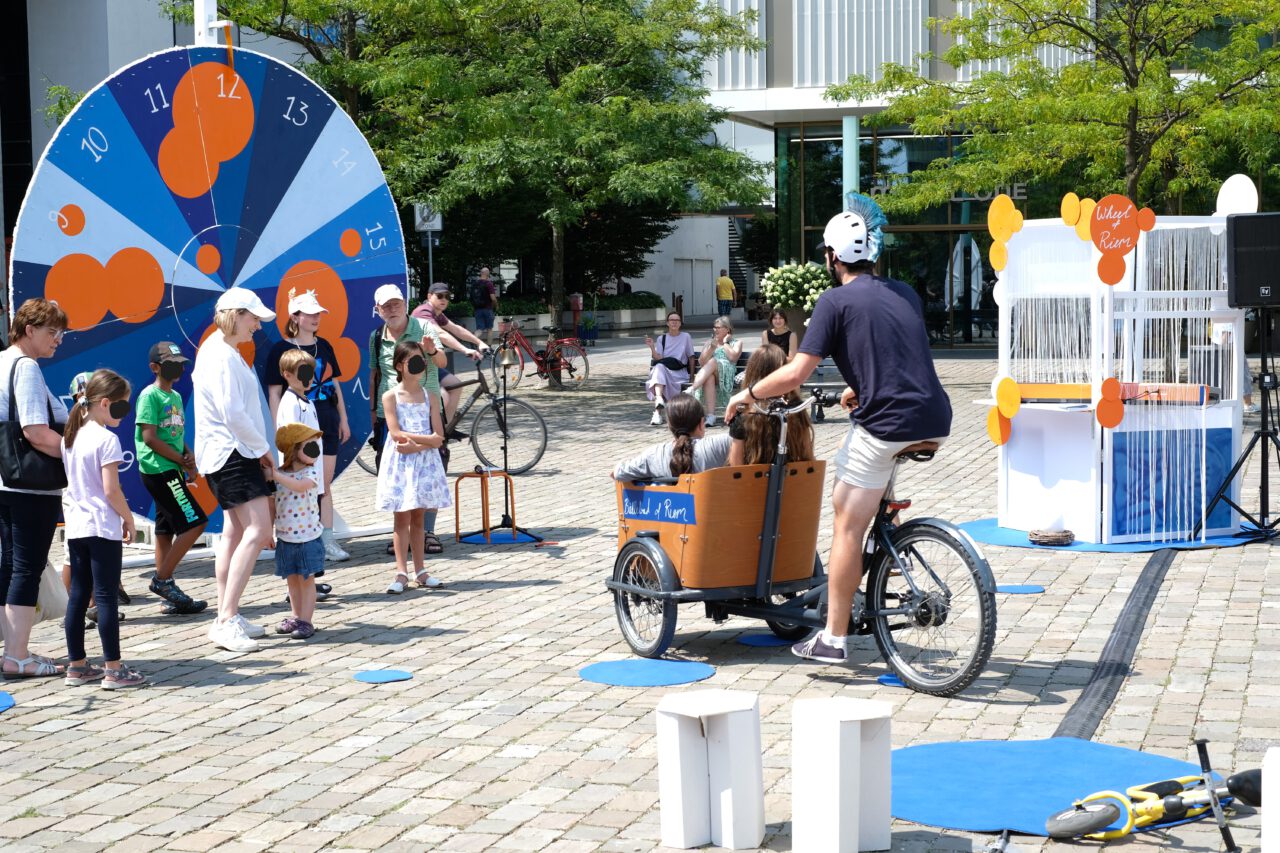 Auf einem öffentlichen Platz findet eine interaktive Performance statt. Eine Gruppe von Erwachsenen und Kindern steht vor einem großen blau-weißen Glücksrad mit orangefarbenen Elementen. Im Vordergrund fährt eine Person mit einem Lastenfahrrad, in dem Kinder sitzen, auf die Gruppe zu. Rechts steht eine Installation mit hängenden Fäden und dem Schild „Wheel of Rhyme“. Die Szene zeigt eine lebendige, sonnige kulturelle Veranstaltung im Freien.