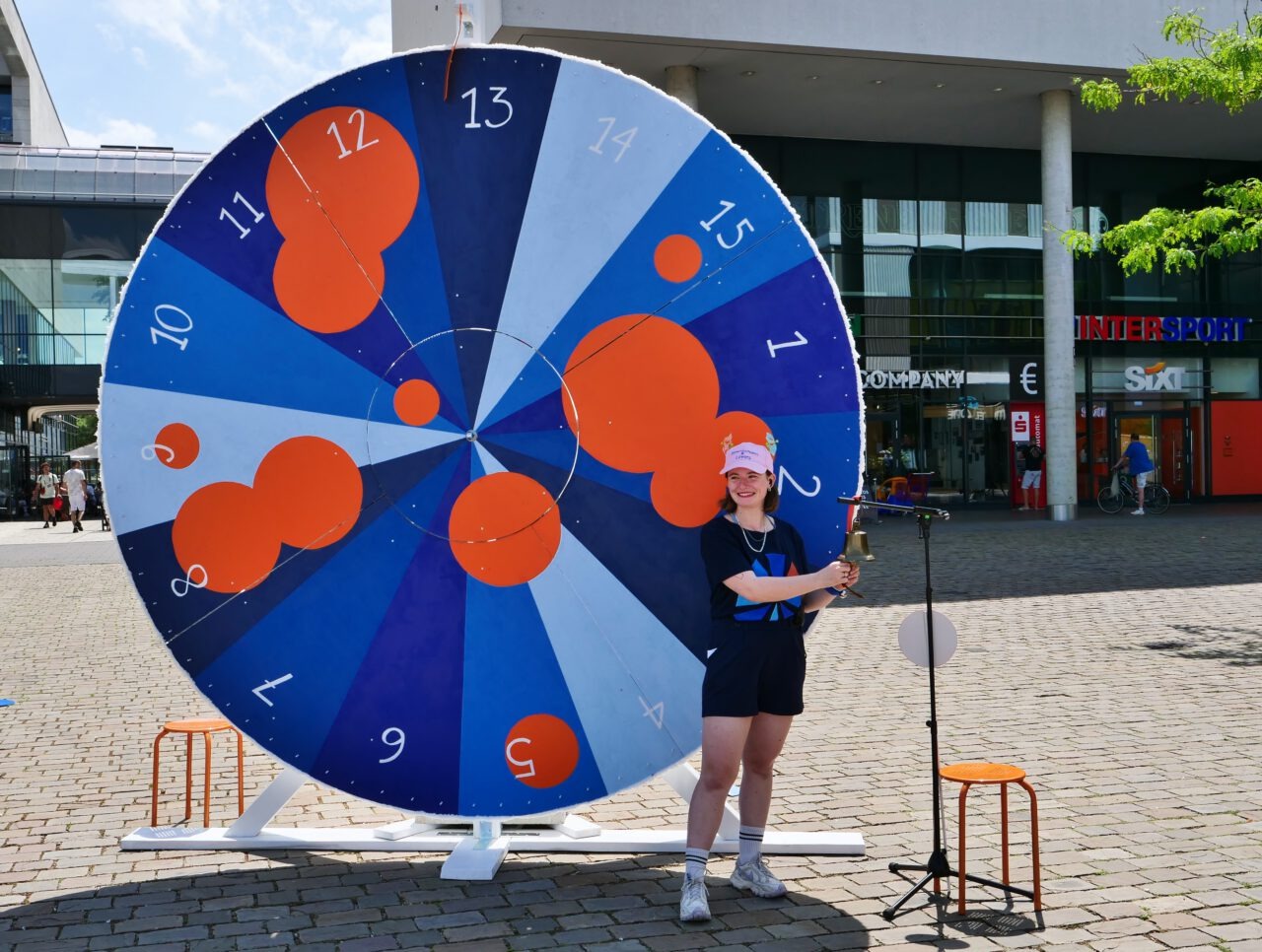 Auf einem großen Platz im Freien steht ein überdimensionales Glücksrad. Das Rad ist in verschiedenen Blau- und Weißtönen gestaltet und in nummerierte Segmente von 1 bis 15 unterteilt. Mehrere große und kleine orangefarbene Kreise sind über die Fläche verteilt. Rechts vor dem Rad steht eine lächelnde Person. Sie trägt dunkle Kleidung, helle Sportschuhe und eine rosafarbene Kappe. In den Händen hält sie eine kleine Glocke. Neben ihr befindet sich ein Mikrofonständer. Links und rechts des Glücksrads stehen jeweils kleine orangefarbene Hocker. Im Hintergrund sind moderne Geschäftsgebäude mit großen Glasflächen zu sehen. Das Foto wurde bei sonnigem Wetter aufgenommen.