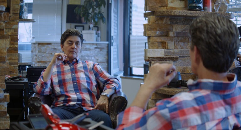 Das Bild zeigt eine Gesprächssituation in einem modernen Friseursalon. Eine Person sitzt entspannt in einem Ledersessel und blickt aufmerksam nach vorn, während sie sich offenbar mit einer anderen Person unterhält, die im Vordergrund nur teilweise von hinten zu sehen ist. Die sitzende Person trägt ein rot-blau kariertes Hemd und wirkt konzentriert und nachdenklich. Die Umgebung ist stilvoll gestaltet: Steinwände, große Fenster mit Jalousien sowie Pflanzen und dekorative Elemente schaffen eine warme, professionelle Atmosphäre. Im Hintergrund sind typische Salon-Elemente zu erkennen, darunter Arbeitsflächen und Friseurwerkzeuge. Das Bild vermittelt einen ruhigen Moment der Beratung oder des Austauschs in einem gepflegten, modernen Umfeld.