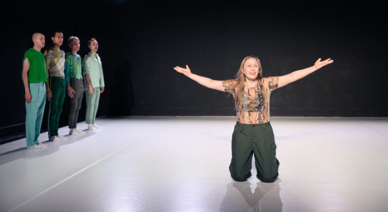 Eine Frau kniet im Zentrum einer minimalistischen Bühne und streckt die Arme weit aus – eine ausdrucksstarke, beinahe triumphale Geste. Mit offenem, strahlendem Gesicht wendet sie sich dem Publikum zu. Im Hintergrund stehen vier weitere Performer*innen in einer Reihe vor einer dunklen Wand, gekleidet in aufeinander abgestimmten Grüntönen. Die helle, weiß ausgeleuchtete Bühnenfläche bildet einen starken Kontrast zur dunklen Umgebung und rückt die zentrale Figur in den Fokus. Die Szene vermittelt eine intensive, theatralische Atmosphäre voller Spannung und emotionaler Offenheit.