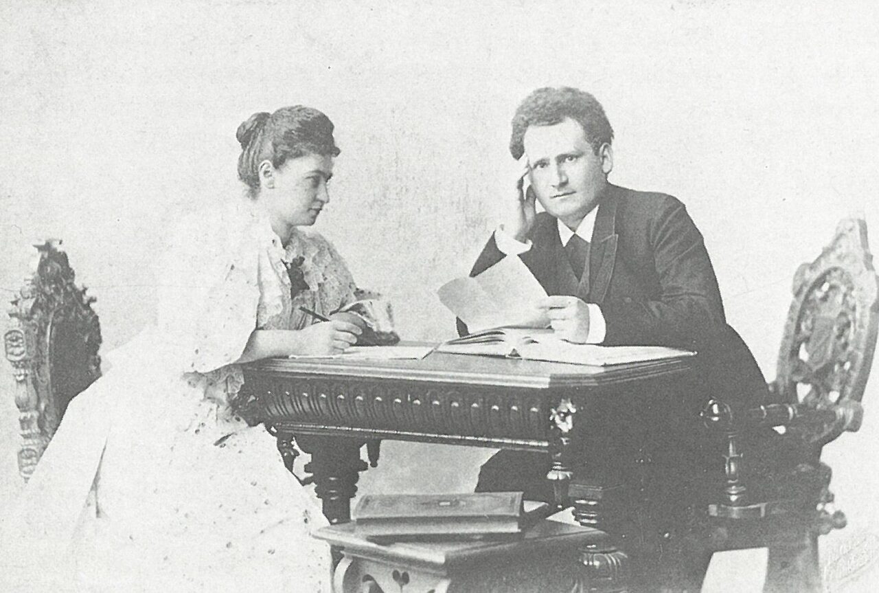 Historische Schwarz-Weiß-Fotografie eines Mannes und einer Frau um 1900, die an einem Tisch sitzen und gemeinsam Dokumente lesen.