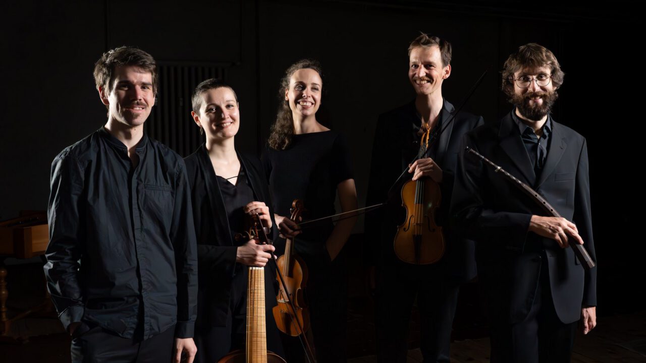 Auf dem Bild ist ein klassisches Musikensemble zu sehen, bestehend aus fünf Musikerinnen und Musikern. Sie stehen nebeneinander in einem dunklen, atmosphärisch beleuchteten Raum und schauen lächelnd in die Kamera. Alle tragen elegante, überwiegend schwarze Kleidung, die einen professionellen und stilvollen Eindruck vermittelt. Einige der Personen halten ihre Instrumente in den Händen: Zu erkennen sind unter anderem eine Violine sowie ein barockes Streichinstrument (ähnlich einer Viola da gamba) und eine Querflöte. Die warme Beleuchtung hebt die Gesichter und Instrumente hervor, während der Hintergrund bewusst dunkel gehalten ist, wodurch die Gruppe besonders im Fokus steht.
