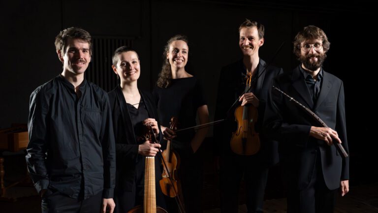 Auf dem Bild ist ein klassisches Musikensemble zu sehen, bestehend aus fünf Musikerinnen und Musikern. Sie stehen nebeneinander in einem dunklen, atmosphärisch beleuchteten Raum und schauen lächelnd in die Kamera. Alle tragen elegante, überwiegend schwarze Kleidung, die einen professionellen und stilvollen Eindruck vermittelt. Einige der Personen halten ihre Instrumente in den Händen: Zu erkennen sind unter anderem eine Violine sowie ein barockes Streichinstrument (ähnlich einer Viola da gamba) und eine Querflöte. Die warme Beleuchtung hebt die Gesichter und Instrumente hervor, während der Hintergrund bewusst dunkel gehalten ist, wodurch die Gruppe besonders im Fokus steht.