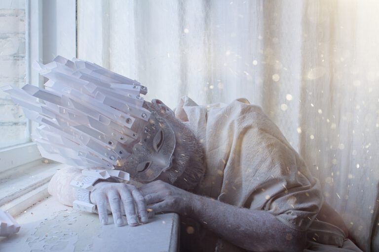 Ein Mensch sitzt am Fenster und hat den Kopf auf die Fensterbank gelegt. Er trägt eine kunstvolle, weiße Maske mit geschlossenen Augen und lange, kantige, kristallartige Elemente, die wie ein zerbrechlicher Kopfschmuck nach vorne ragen. Sein Gesicht und seine Hände sind weiß geschminkt, die Kleidung ist hell und schlicht. Das weiche Tageslicht fällt von links durch das Fenster und taucht die Szene in eine kühle, fast frostige Stimmung. Kleine helle Partikel schweben in der Luft und verleihen dem Bild eine ruhige, verträumte und etwas melancholische Atmosphäre.