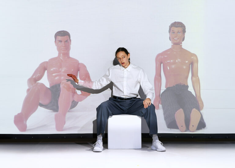 Person in a white shirt and grey trousers sits on a white block in front of a projection showing two large male figurines.