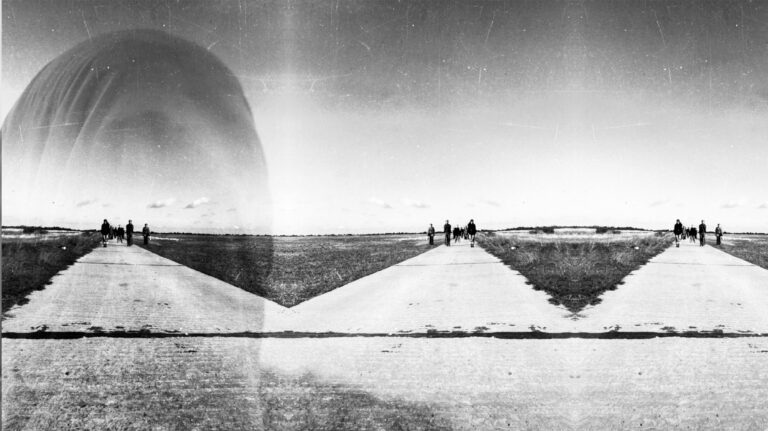 Schwarz-Weiß-Fotografie einer weitläufigen, flachen Landschaft. Mehrere Gruppen von Menschen gehen auf hellen, geraden Wegen, die sich fächerförmig durch eine offene, grasbewachsene Fläche ziehen. Im Hintergrund erstreckt sich eine niedrige Horizontlinie unter einem hellen Himmel. Über der Landschaft ist schemenhaft und halbtransparent das Gesicht einer Person eingeblendet, wodurch ein Überblendungs- oder Doppelbelichtungseffekt entsteht. Die historische Aufnahme weist sichtbare Kratzer und Gebrauchsspuren auf.