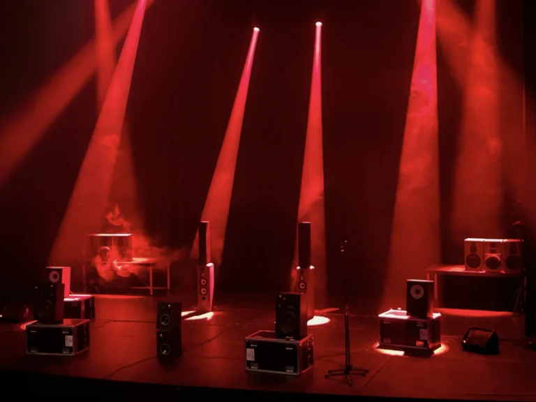 Auf einer dunklen Bühne stehen mehrere Lautsprecher und Audiogeräte verteilt auf dem Boden und auf kleinen Tischen. Die Szene ist in intensives rotes Licht getaucht. Von der Decke fallen mehrere schmale, helle Lichtstrahlen nach unten und beleuchten einzelne Bereiche der Bühne. In der Mitte steht ein Mikrofonständer. Um ihn herum sind verschiedene Lautsprecherboxen in unterschiedlichen Größen angeordnet, einige stehen auf Transportkisten. Links auf einem Tisch befindet sich weiteres Audioequipment hinter einer transparenten Schutzhaube, aus der leichter Nebel oder Rauch aufsteigt. Auch auf der rechten Seite steht ein Tisch mit Lautsprechern.