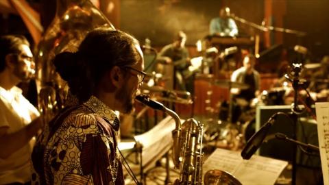 Eine Person mit zusammengebundenen Haaren und Brille spielt im Vordergrund Saxofon und sitzt vor einem Notenständer mit aufgelegten Noten. Im Hintergrund sind weitere Musikerinnen und Musiker mit verschiedenen Instrumenten, darunter Schlagzeug und Blechblasinstrumente, zu sehen. Die Szene ist in warmes, goldfarbenes Bühnenlicht getaucht und vermittelt die konzentrierte Atmosphäre einer Musikprobe oder eines Konzerts. Der Fokus liegt auf der saxofonspielenden Person im Vordergrund, während der Hintergrund leicht unscharf erscheint.