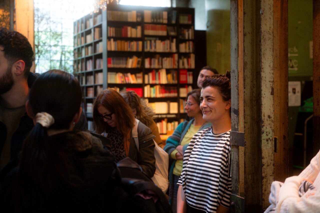 Menschen stehen in einer Bücherei-Atmosphäre, eine Frau im gestreiften Shirt lächelt, während andere zuhören oder beschäftigt sind.