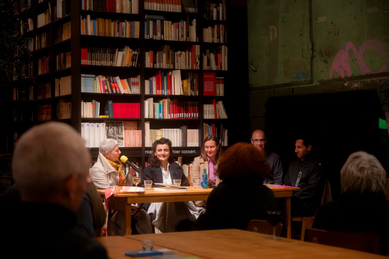 Podiumsdiskussion vor einer Bücherwand, fünf Personen sitzen an einem Tisch und sprechen vor Publikum.