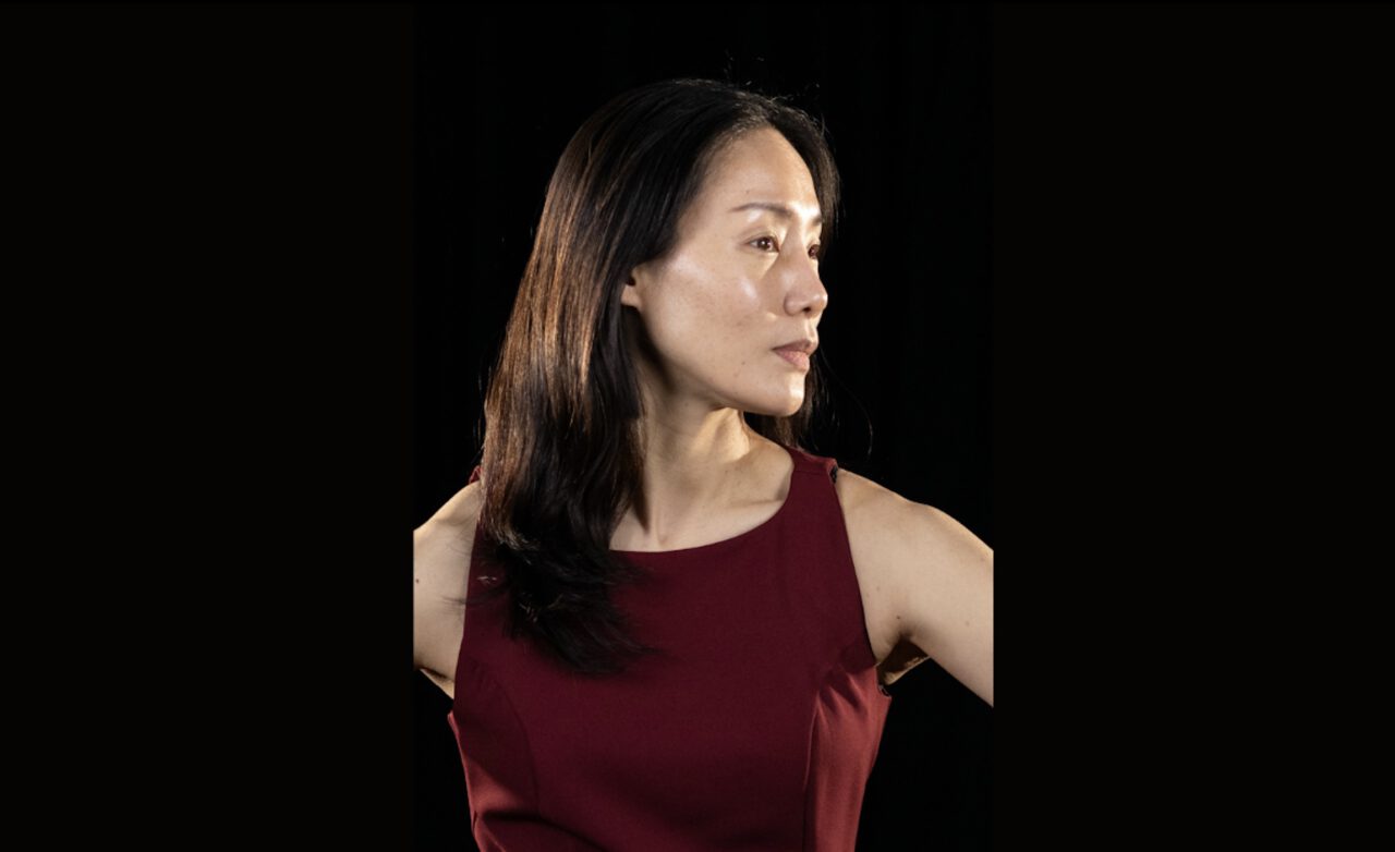Portrait of a person against a black background. The person is wearing a sleeveless burgundy top and has shoulder-length dark hair. Their upper body faces the camera, while their head is turned to the right and their gaze is directed into the distance. Warm side lighting illuminates the face, highlighting its contours and skin texture. The arms are bent outward at the sides and are partially visible in the frame.